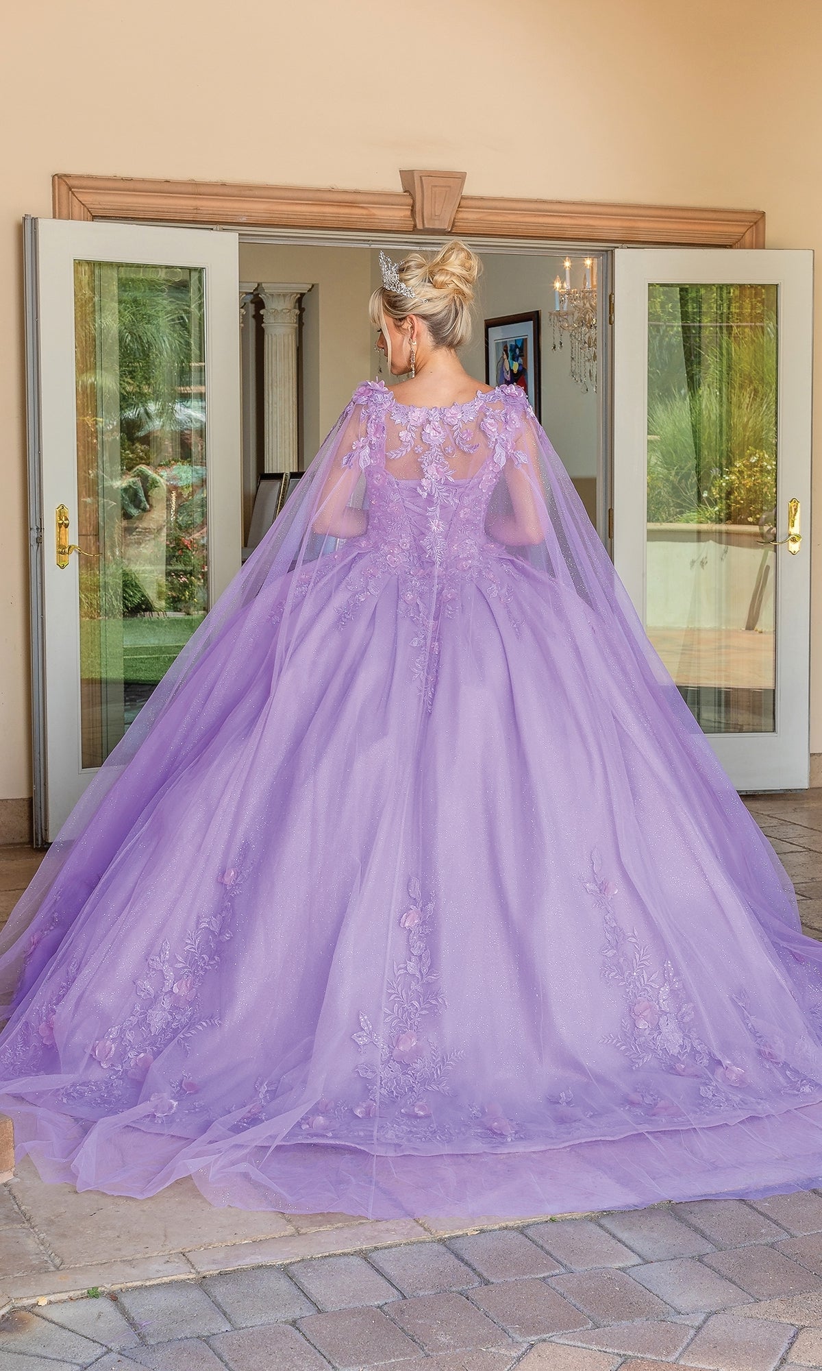 A woman stands facing away in front of open double doors, wearing the Quinceanera Dress 1716 by Dancing Queen—a sleeveless ball gown with floral embroidery, a cape, and a sweetheart neckline.